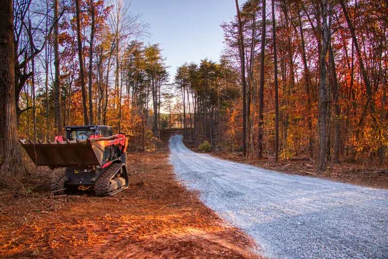 Compact skid steer parked beside a freshly cleared and gravelled rural driveway through autumn forest
